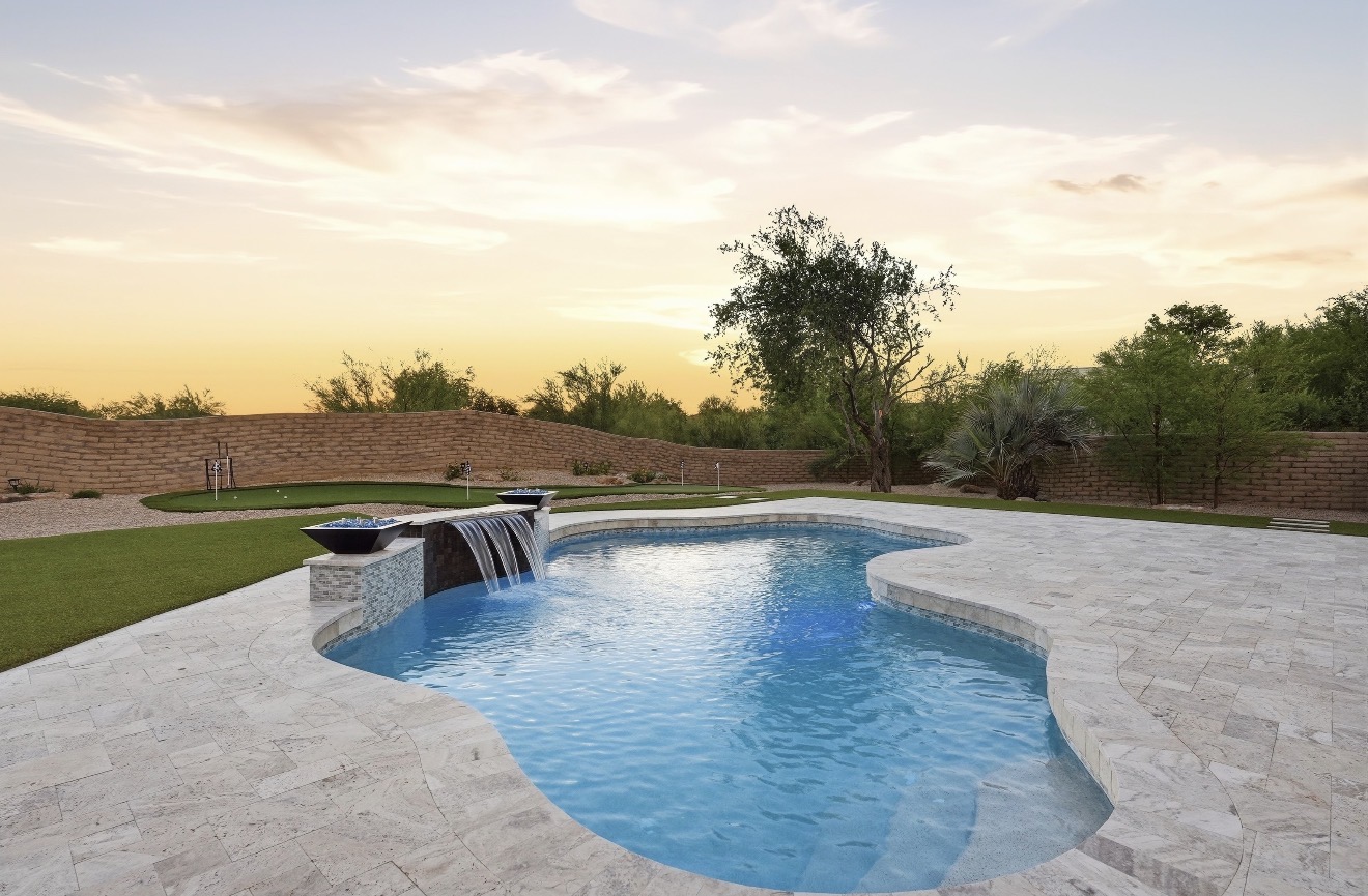 A freeform backyard pool at golden hour with a sheer waterfall, fire bowls, and a putting green beyond the travertine deck