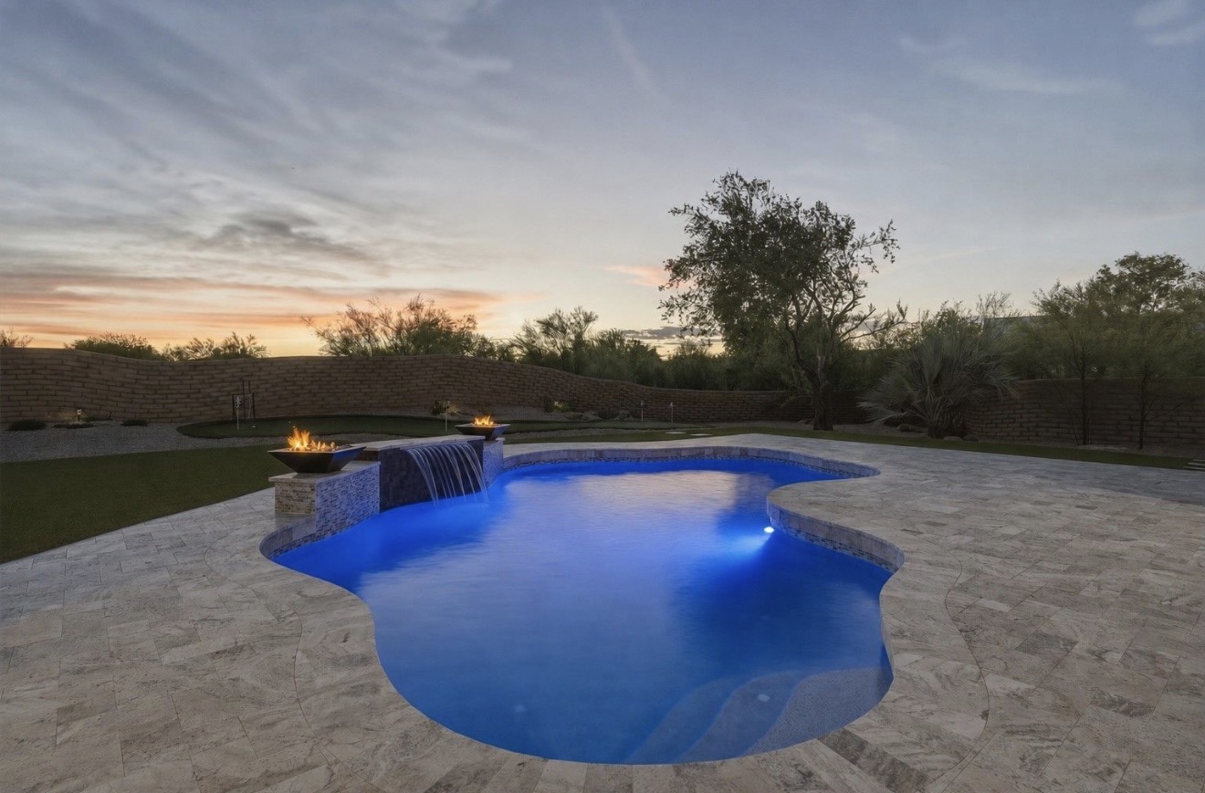 Freeform backyard pool lit blue at dusk with a sheer waterfall and fire bowls